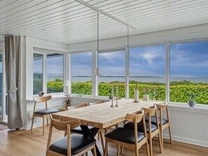 Un vaste espace dédié à la salle à manger, avec une grande table et des chaises, de grandes fenêtres offrant une vue sur la verdure et l’eau, et un plafond blanc avec des poutres apparentes.