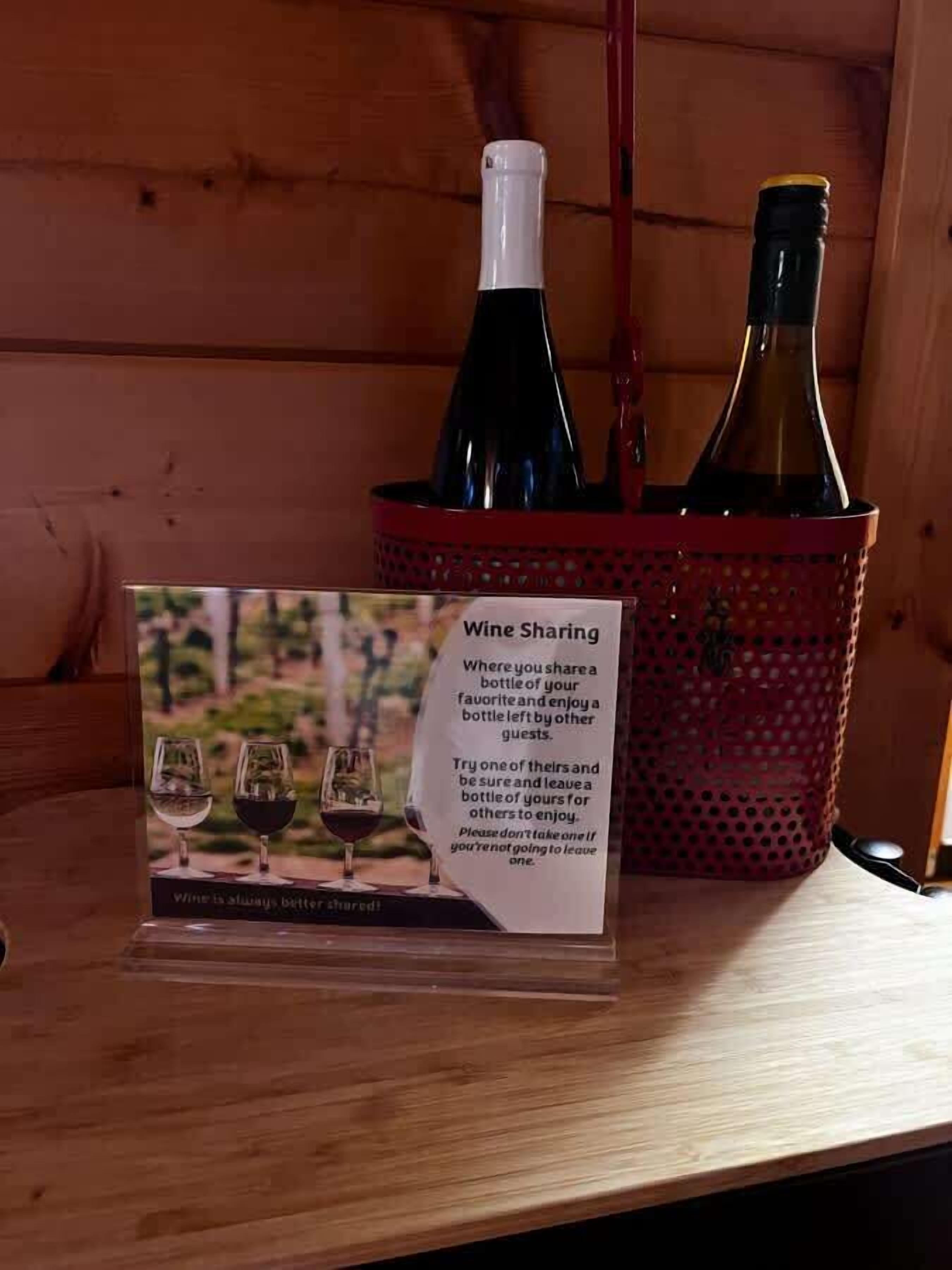 Un panneau indiquant qu’il est possible de partager du vin, accompagné d’instructions, placé à côté d’une table en bois sur laquelle se trouvent deux bouteilles de vin dans un panier rouge.