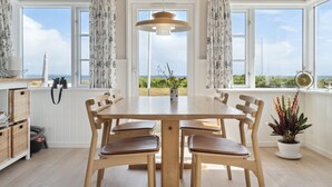 Uma área de refeições com mesa e cadeiras de madeira, grandes janelas com cortinas, luminária pendente e vista para o mar.