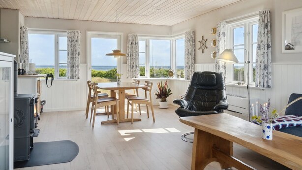 Um quarto acolhedor com teto de madeira, uma mesa de jantar com cadeiras, uma poltrona de couro preto, uma escrivaninha e vista para o mar através de grandes janelas.