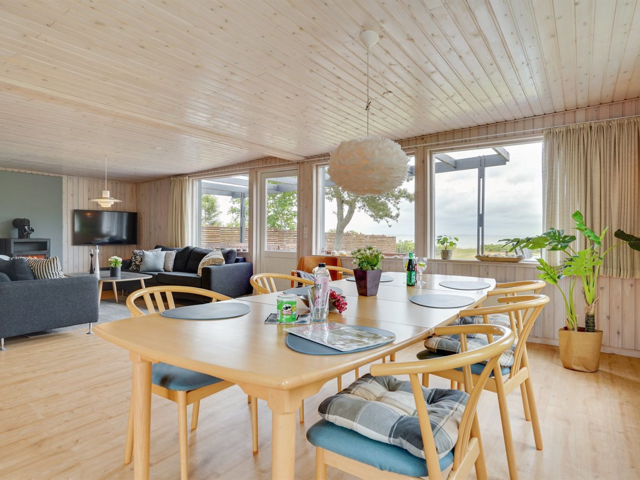 Una sala de estar moderna con techo de madera, televisor grande, sofá, mesa de comedor con sillas y vistas al exterior.