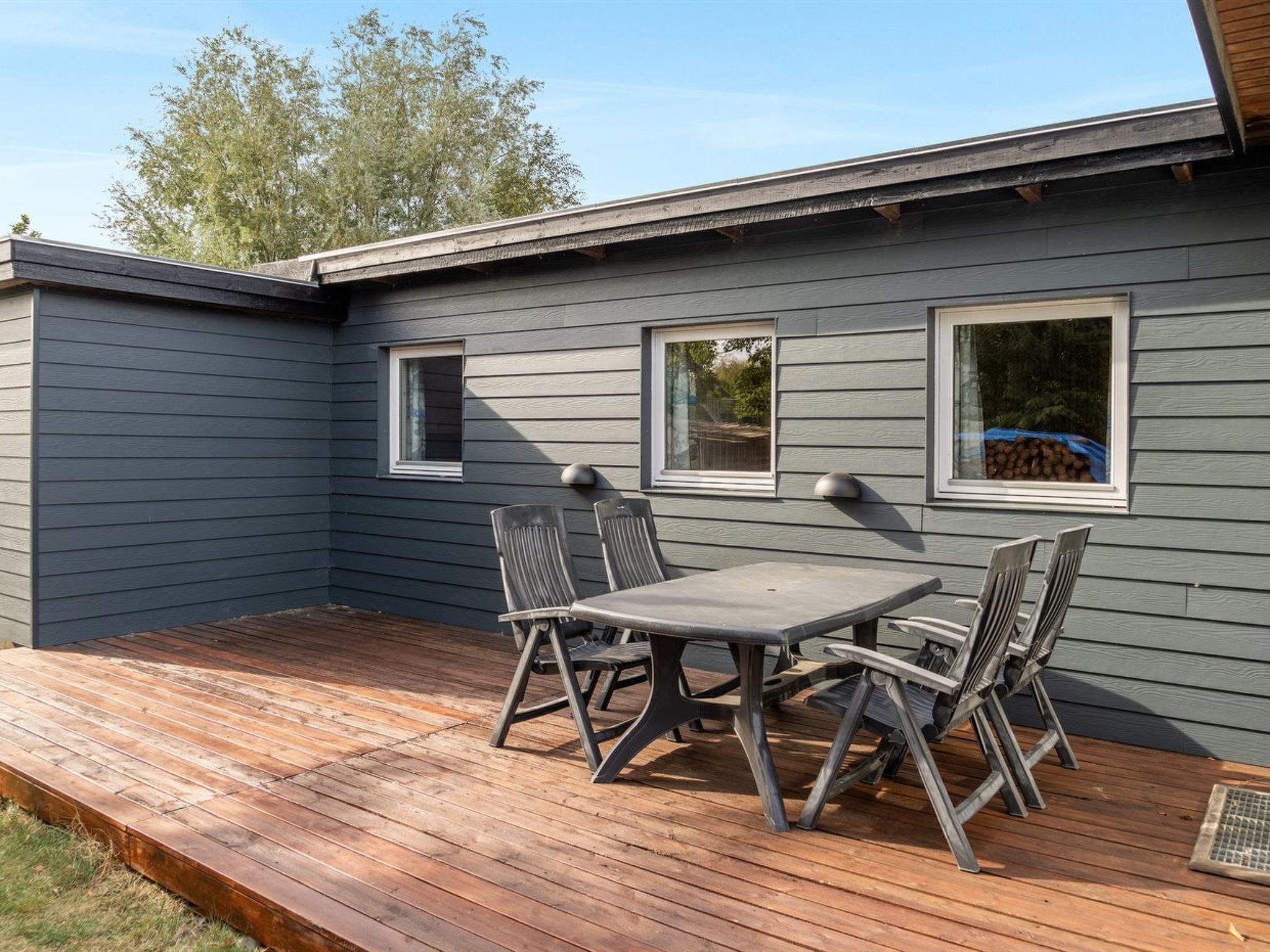 A covered patio with a table and chairs, wooden deck, and a house with grey siding and windows.