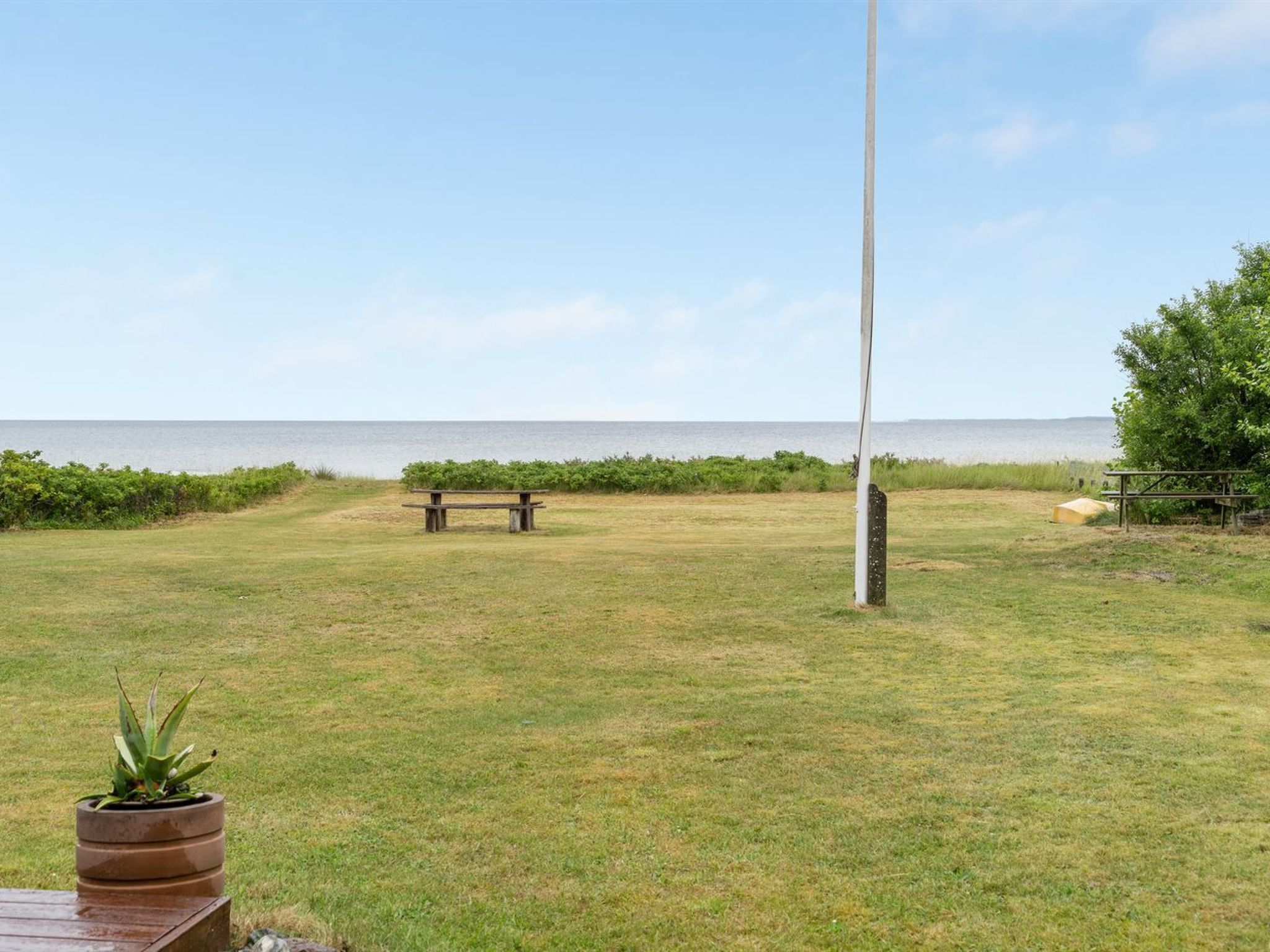 An outdoor area with a grassy field, a wooden bench, a tall flagpole, and a view of the sea.