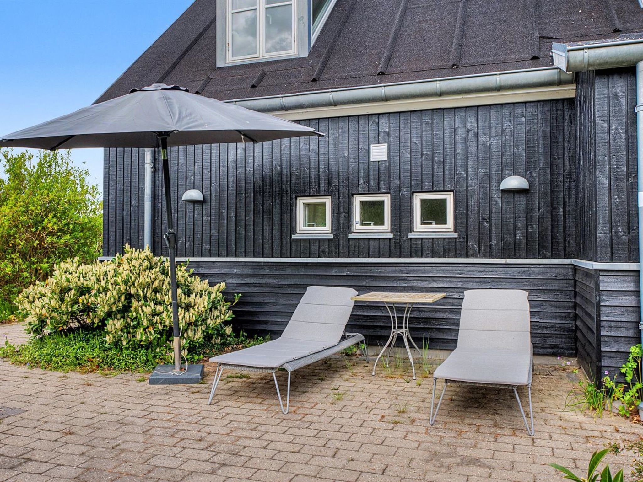 A building with a dark wooden exterior, three windows, and a patio area with two lounge chairs and a table.
