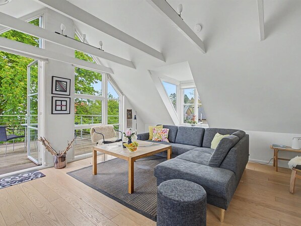 Un loft espacioso con un sofá grande de color gris, una mesa de centro de madera y vistas a la vegetación.