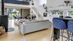 En rummelig opholdsstue med en grå sofa, et spisebord med stole, et køkken med hvide skabe og en trappe til en etage over.