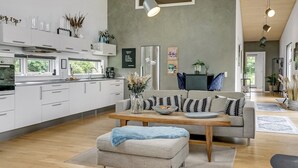 Et moderne køkken med hvide skabe, et spiseområde med en grå sofa og skammel, og en stue med et sofabord.