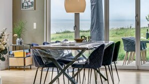 Et spiseområde med et bord, stole og havudsigt gennem store vinduer.