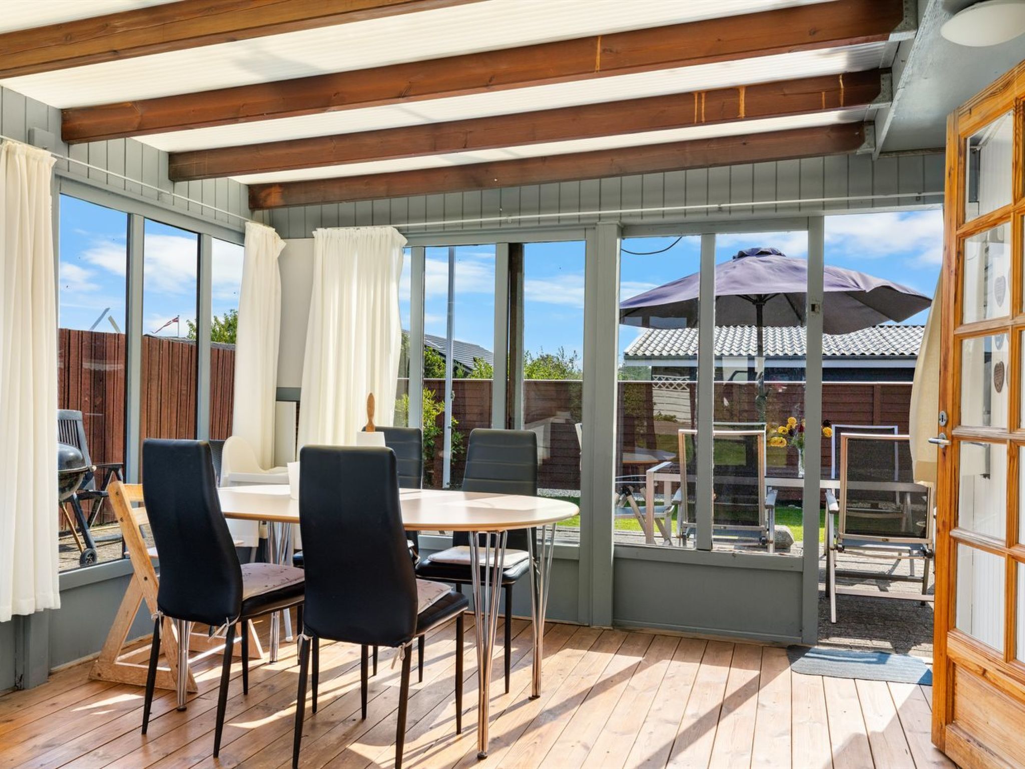 Eine überdachte Terrasse mit Holztisch und -stühlen, großen Fenstern und Blick auf den Garten.