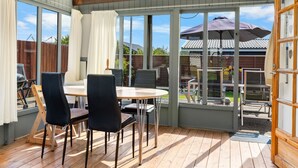 Een overdekt terras met een houten tafel en stoelen, grote ramen en uitzicht op de achtertuin.