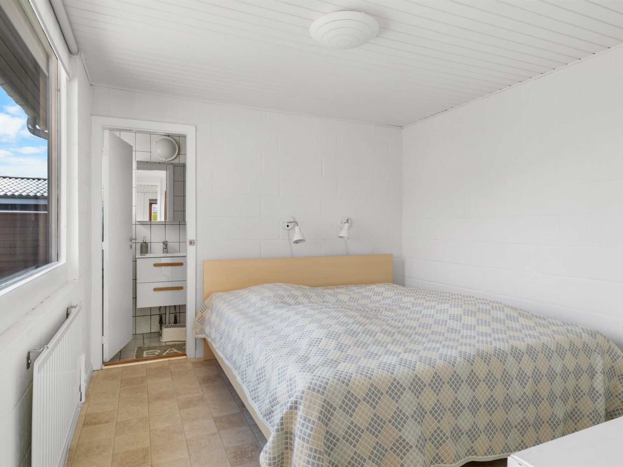 A bedroom with a bed, a nightstand, a window, and a bathroom visible through an open door.