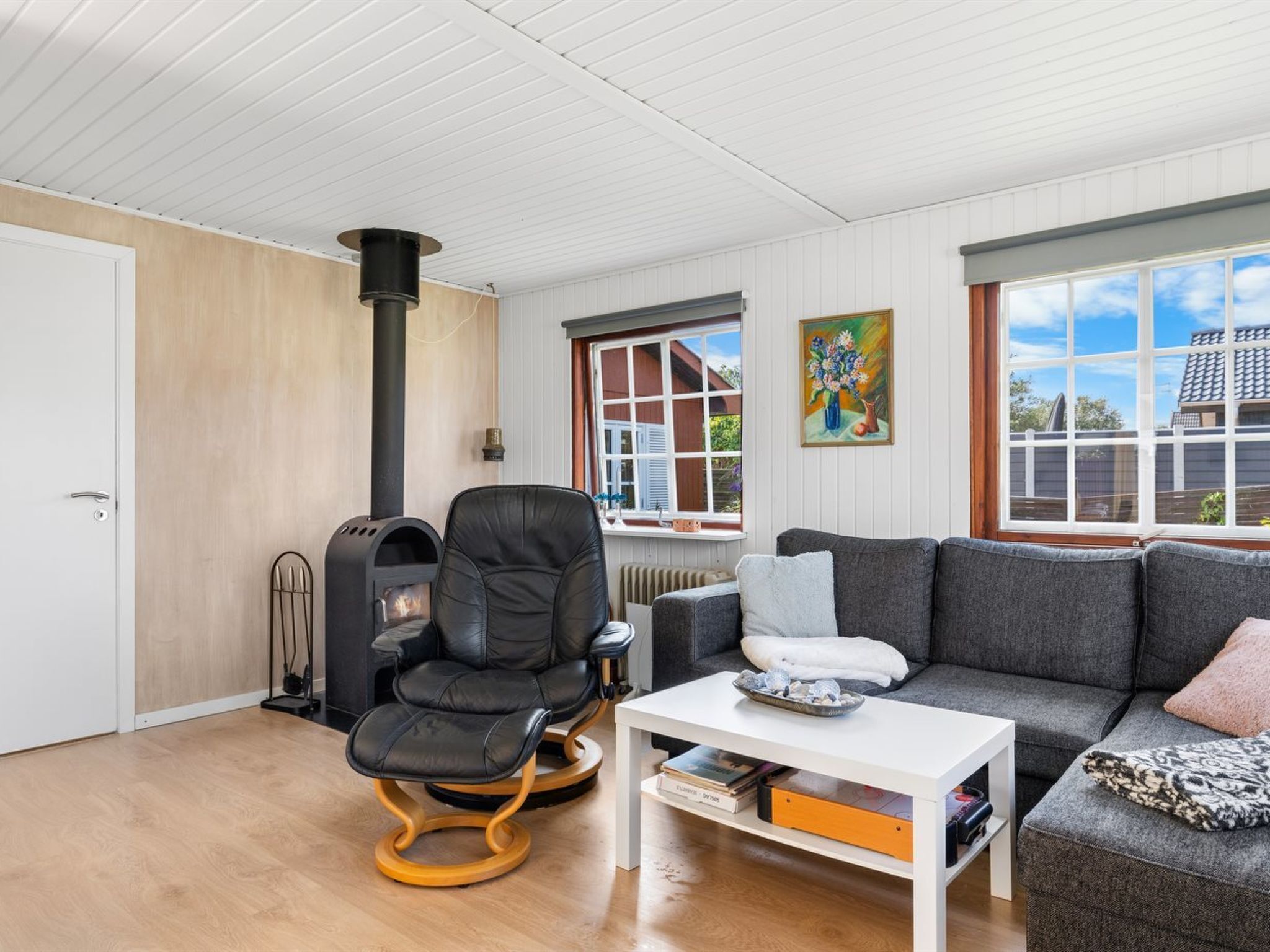 Ein gemütliches Wohnzimmer mit einem schwarzen Ledersessel, einem weißen Couchtisch und einem grauen Sofa. In der Ecke steht ein schwarzer Kaminofen.