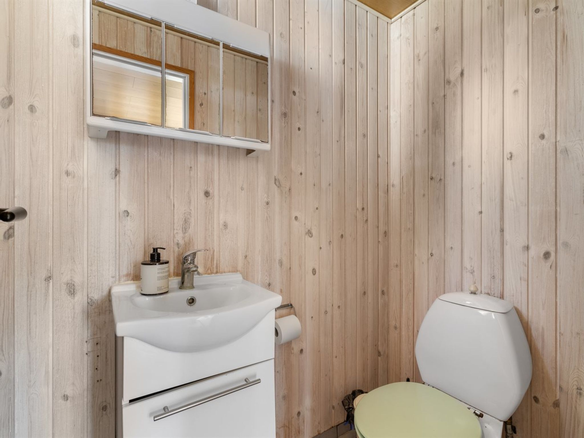 A bathroom with a wooden wall, a white sink with a mirror above it, and a toilet with a green seat.