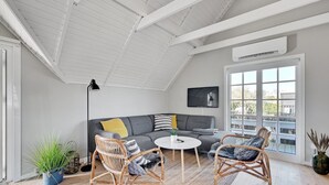 Ein modernes Wohnzimmer mit einem grauen Sofa, zwei Korbstühlen, einem kleinen runden Tisch und einem Fernseher an der Wand.