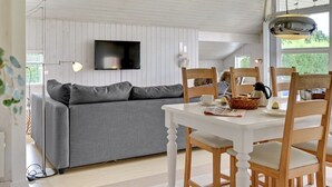 A modern living room with a grey sofa, a white dining table with wooden chairs, a flat-screen TV mounted on the wall, and a large window providing natural light.