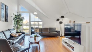 Een ruime woonkamer met een groot raam, een zwarte leren bank, een salontafel en een flatscreen televisie.