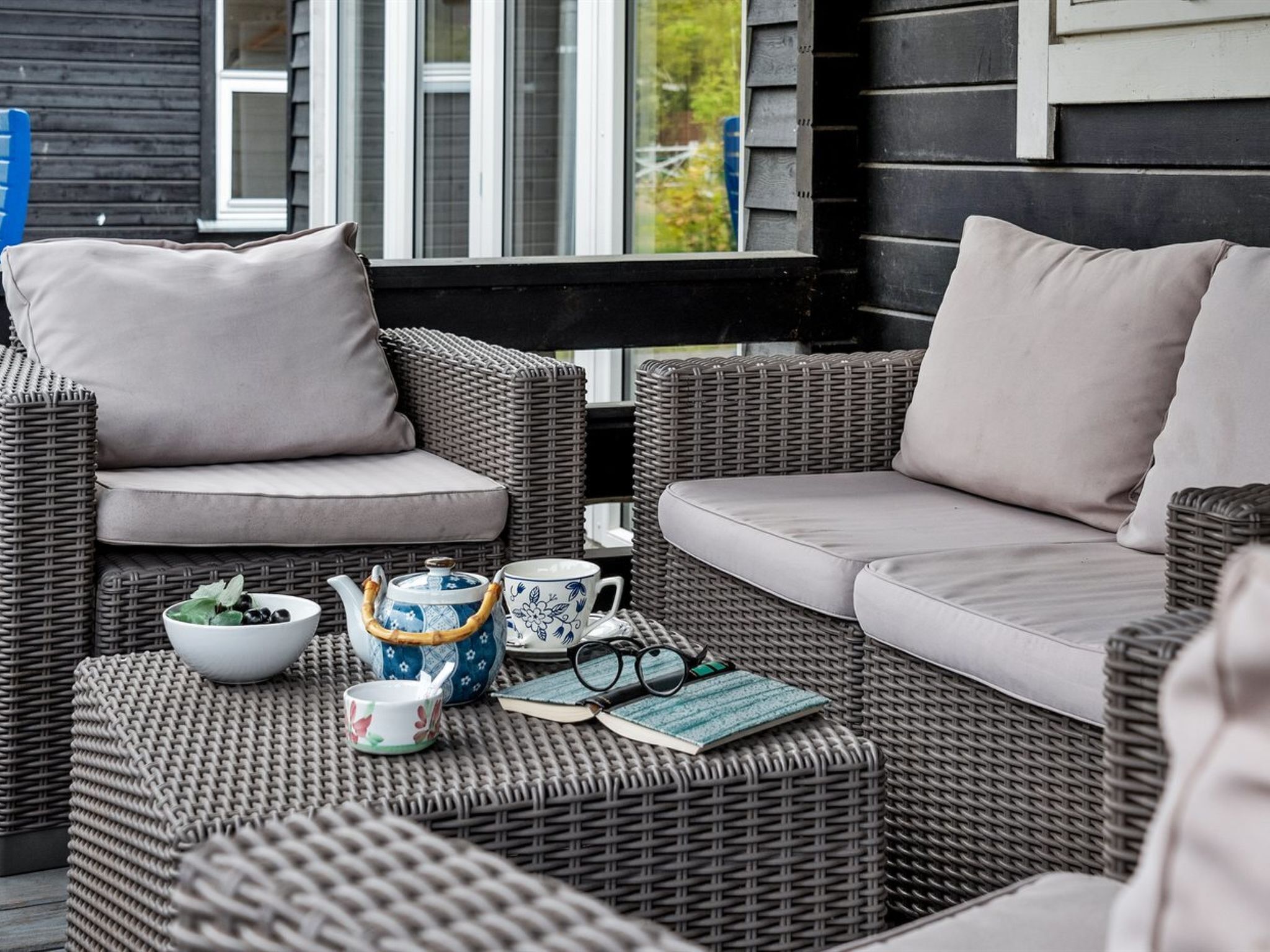 A wicker outdoor seating area with cushions, a table with a teapot, cups, and a book.