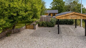 Estructura de madera con la etiqueta "Fruhuset" en un jardín con un sendero de grava, una valla de madera y una casa al fondo.