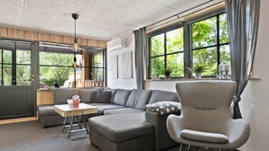 Un salón moderno con un sofá seccional, una mesita y una silla. Hay ventanas amplias con cortinas, un ventilador de techo y vistas a zonas verdes.