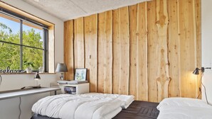Un dormitorio con pared de madera, cama con ropa de cama blanca y negra, escritorio con lámpara y ventana con vista a árboles.