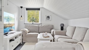 En moderne stue indrettet i hvidt med skråvægge, et stort vindue med udsigt til grønne omgivelser, et fladskærms-tv, en sofa, et sofabord og et sengebord.