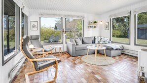 En rummelig stue med en grå sofa, to lænestole, et sofabord og et stort vindue med udsigt over naturen.
