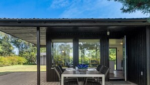 En overdækket terrasse med et bord og stole, store vinduer og udsigt over det grønne område.
