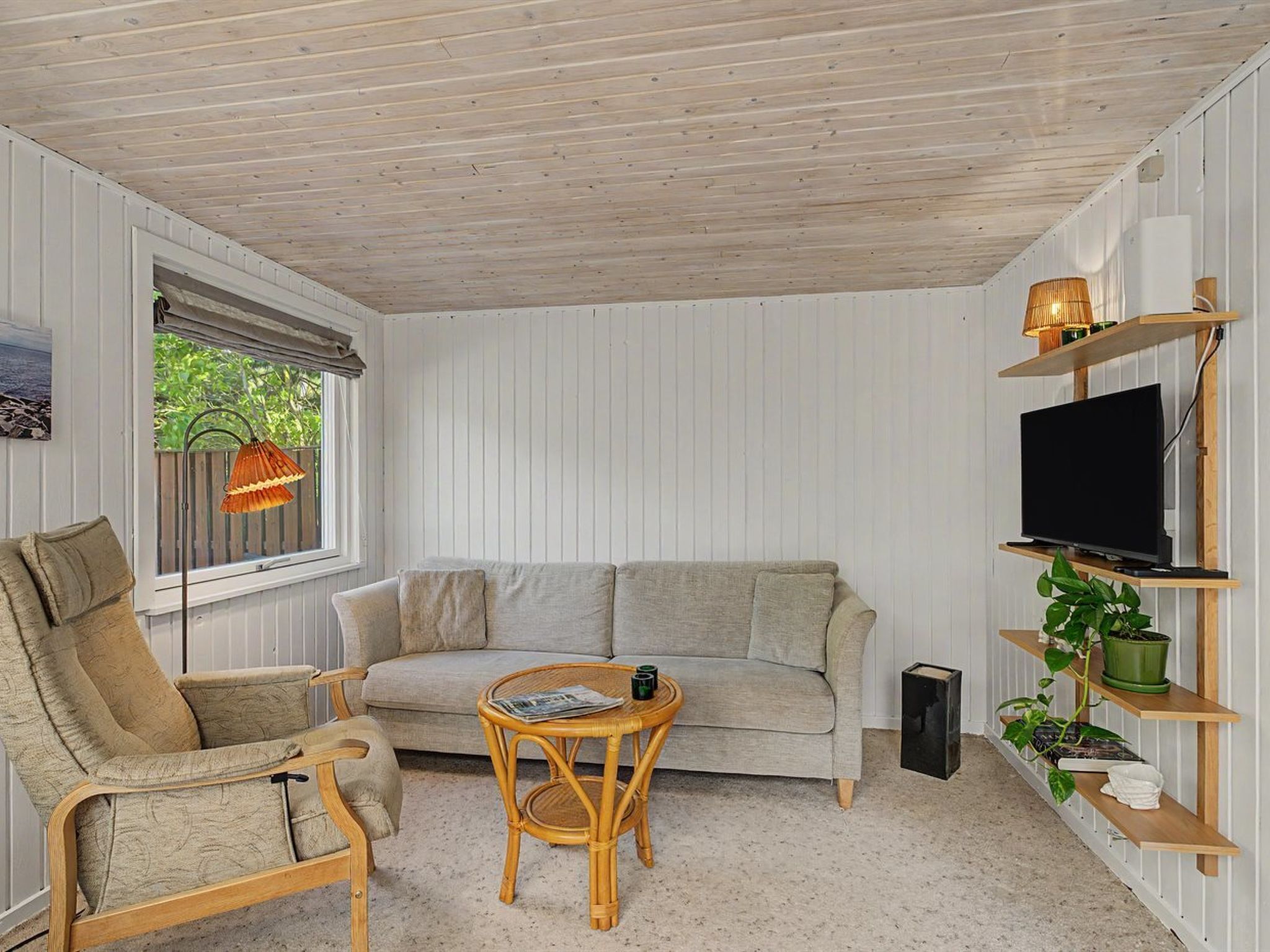 A cozy room with a sofa, armchair, and a small table. There's a TV on a shelf and a window with a view of greenery.