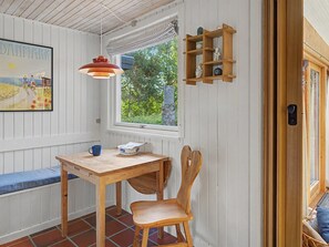 Una habitación con una mesa de madera, un banco, una silla, un ventanal con vistas a la vegetación y un estante en la pared con objetos decorativos.