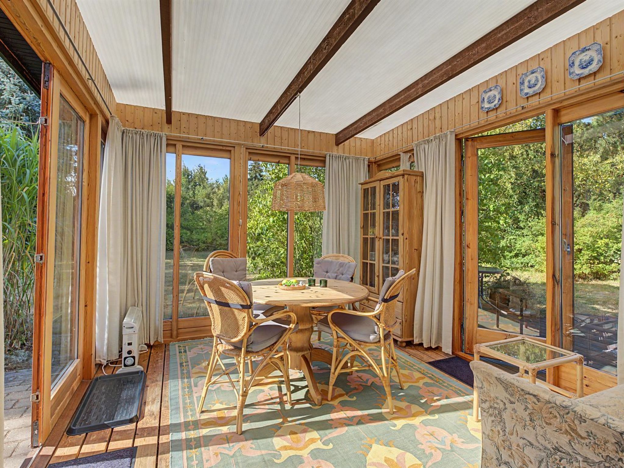 A wooden-paneled room with a round table and chairs, large windows, and a view of greenery outside.