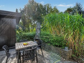 Un patio al aire libre con una mesa y sillas de madera, rodeado de abundante vegetación y un muro de piedra.