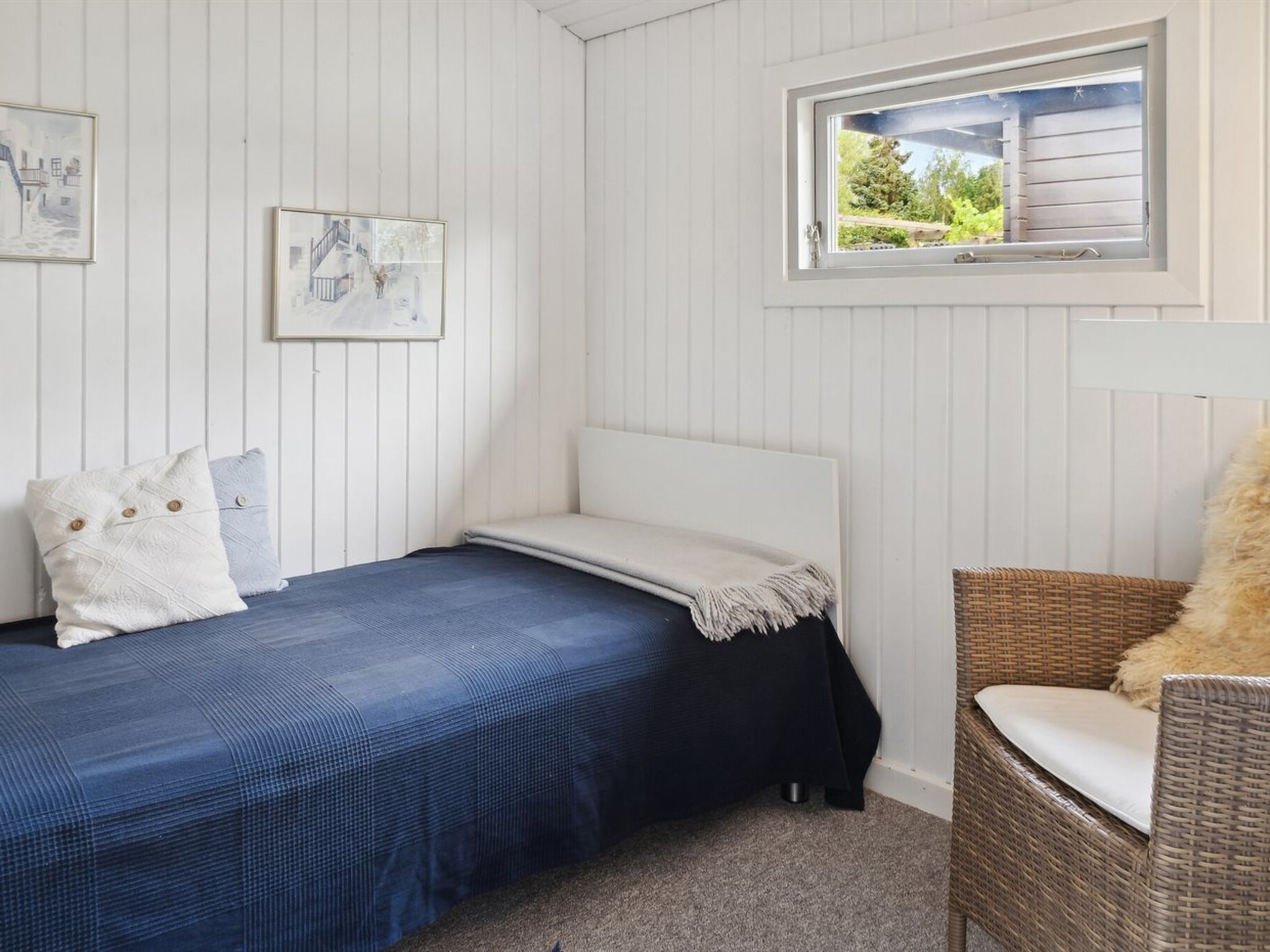 A small, neatly arranged bedroom with a single bed, a window with a view of greenery, and a wicker chair.