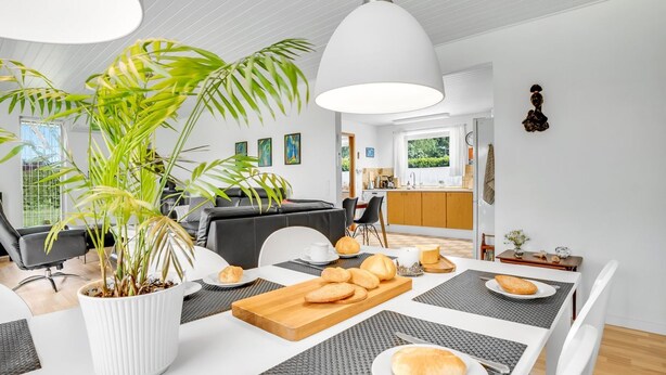A modern kitchen with a dining area, featuring a large potted plant, a wooden cutting board with bread, and a white dining table with grey placemats. - "Maleen" - 150m to the fjord by Interhome (Nakskov)