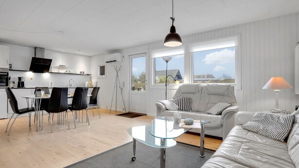 Un salon moderne et bien éclairé, avec une table basse en verre, un canapé et un coin repas comprenant des chaises, et une cuisine en arrière-plan.