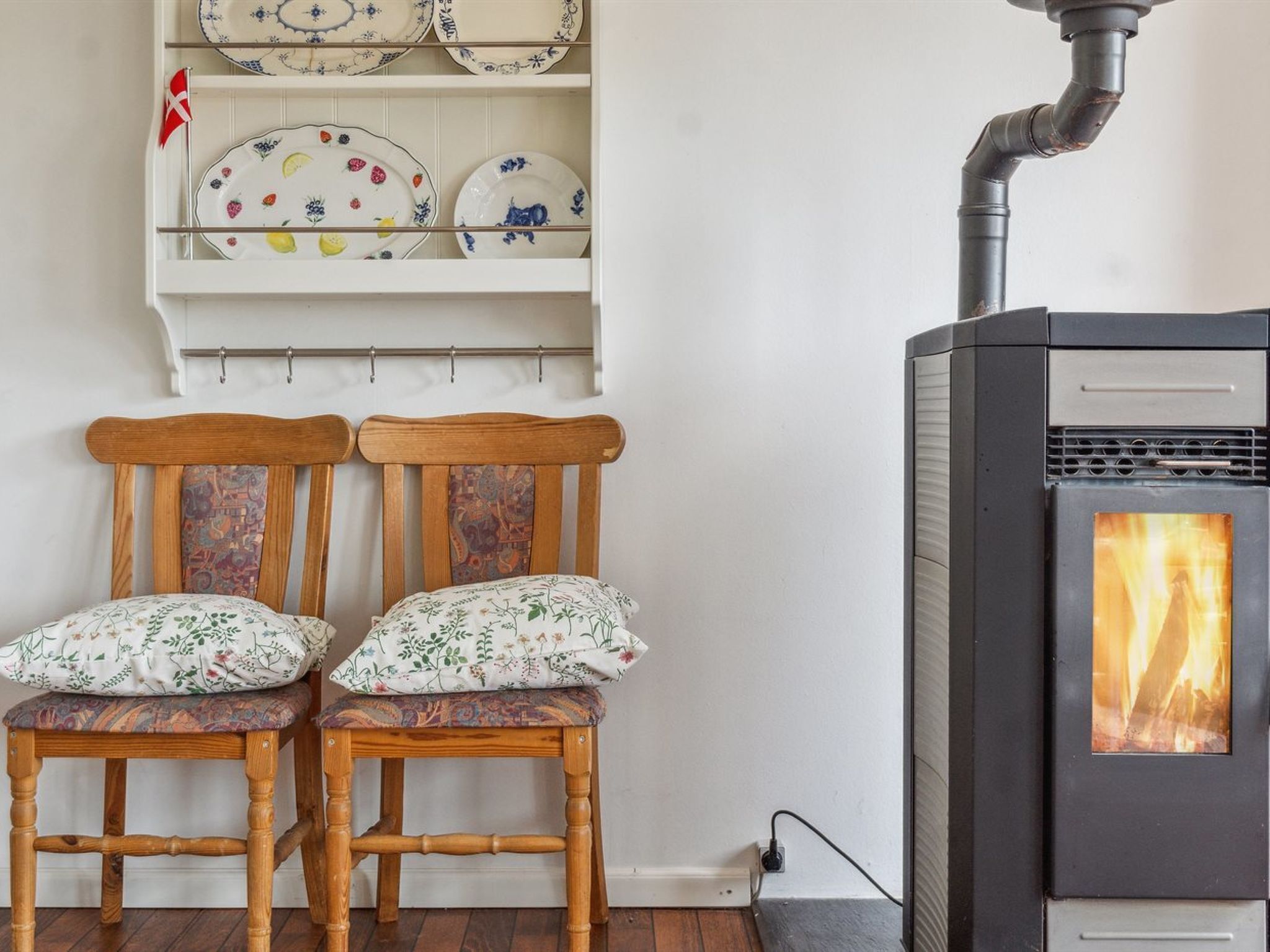 A room with two wooden chairs with floral cushions, a white shelf with decorative plates, and a wood-burning stove.