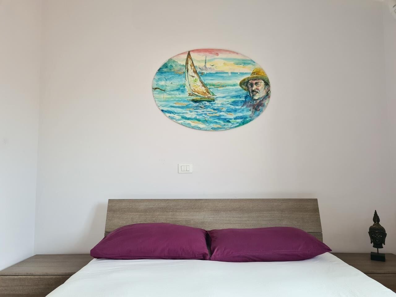 Un lit avec une tête de lit, un mur orné d’un tableau circulaire représentant un voilier, et une petite statue posée sur une table de chevet.