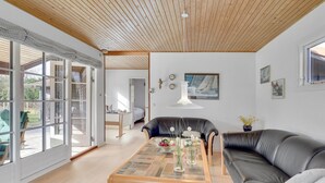 A modern living room with a wooden ceiling, a black leather sofa, a glass coffee table with decorative items, and a view through large glass doors to an outdoor area.