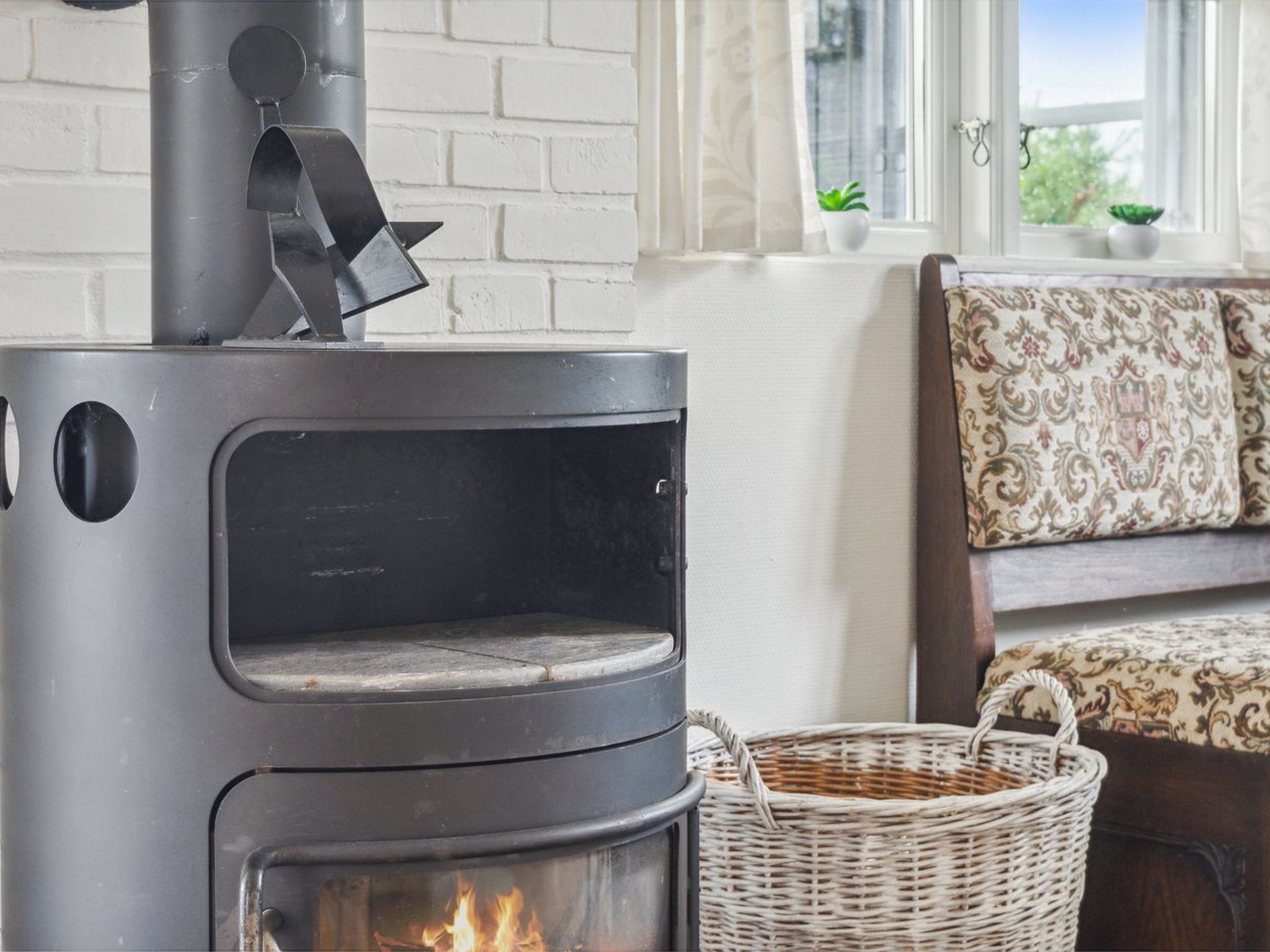 A modern wood-burning stove with a glass door, a wicker basket, and a patterned sofa in a bright room with a white brick wall and a window.