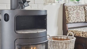 Un poêle à bois moderne avec une porte en verre, un panier en osier et un canapé à motifs dans une pièce lumineuse, dotée d’un mur en briques blanches et d’une fenêtre.