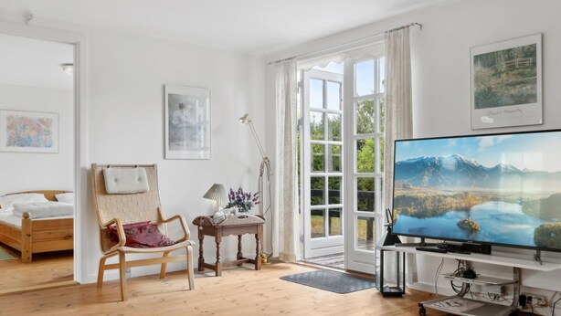 Une pièce spacieuse avec un plancher en bois, un lit, une chaise, un bureau, un téléviseur et une grande fenêtre avec des rideaux.
