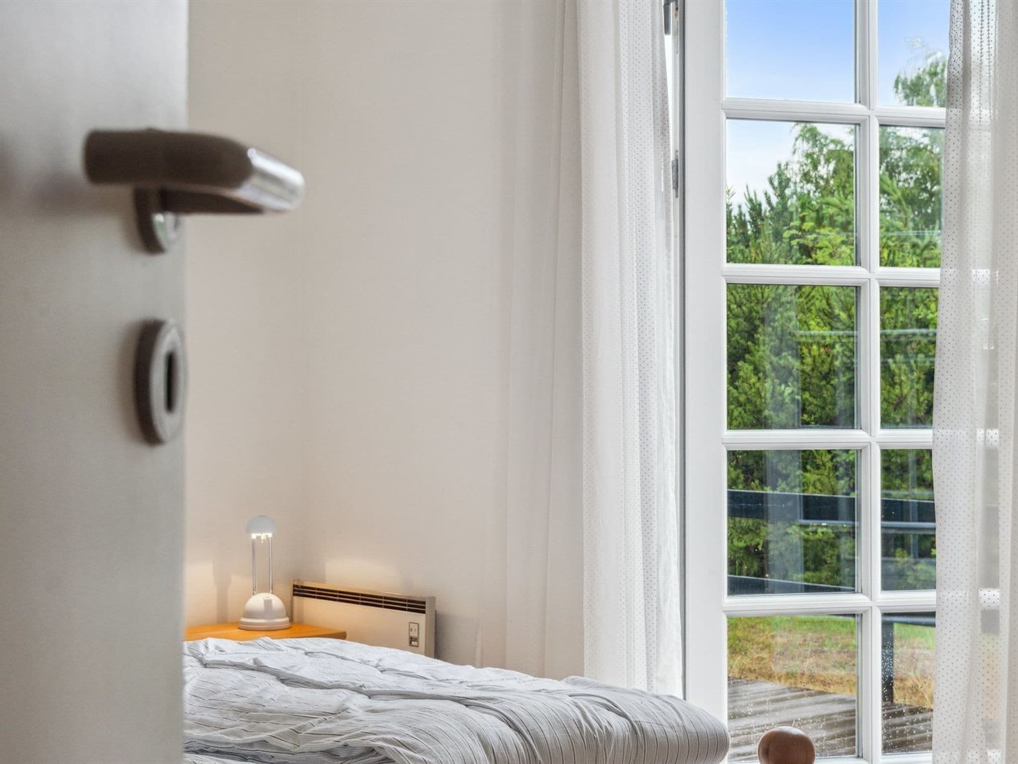 A bedroom with a large window, a bed, a bedside table with a lamp, and a door.
