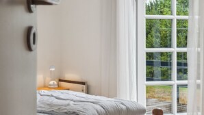 Une chambre avec une grande fenêtre, un lit, une table de chevet avec une lampe et une porte.