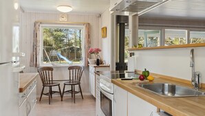 Cocina compacta con área de comedor, sillas de madera, ventana con vista a un tobogán y lavamanos con verduras en la encimera.