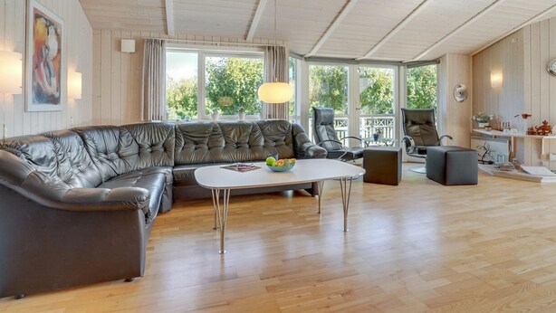 A spacious living room with a leather sectional sofa, a round coffee table, and a dining area with a table and chairs. Large windows provide natural light and a view of greenery outside.