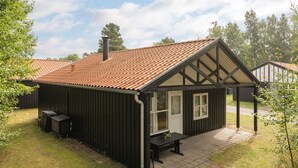 A small, single-story building with a sloped roof and a chimney. It has a porch with a bench and a table, and a small window.