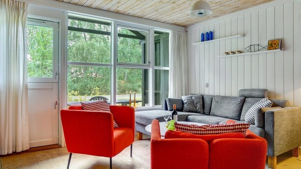 A modern living room with a wooden ceiling, large windows, a grey sofa, red chairs, and a white coffee table.