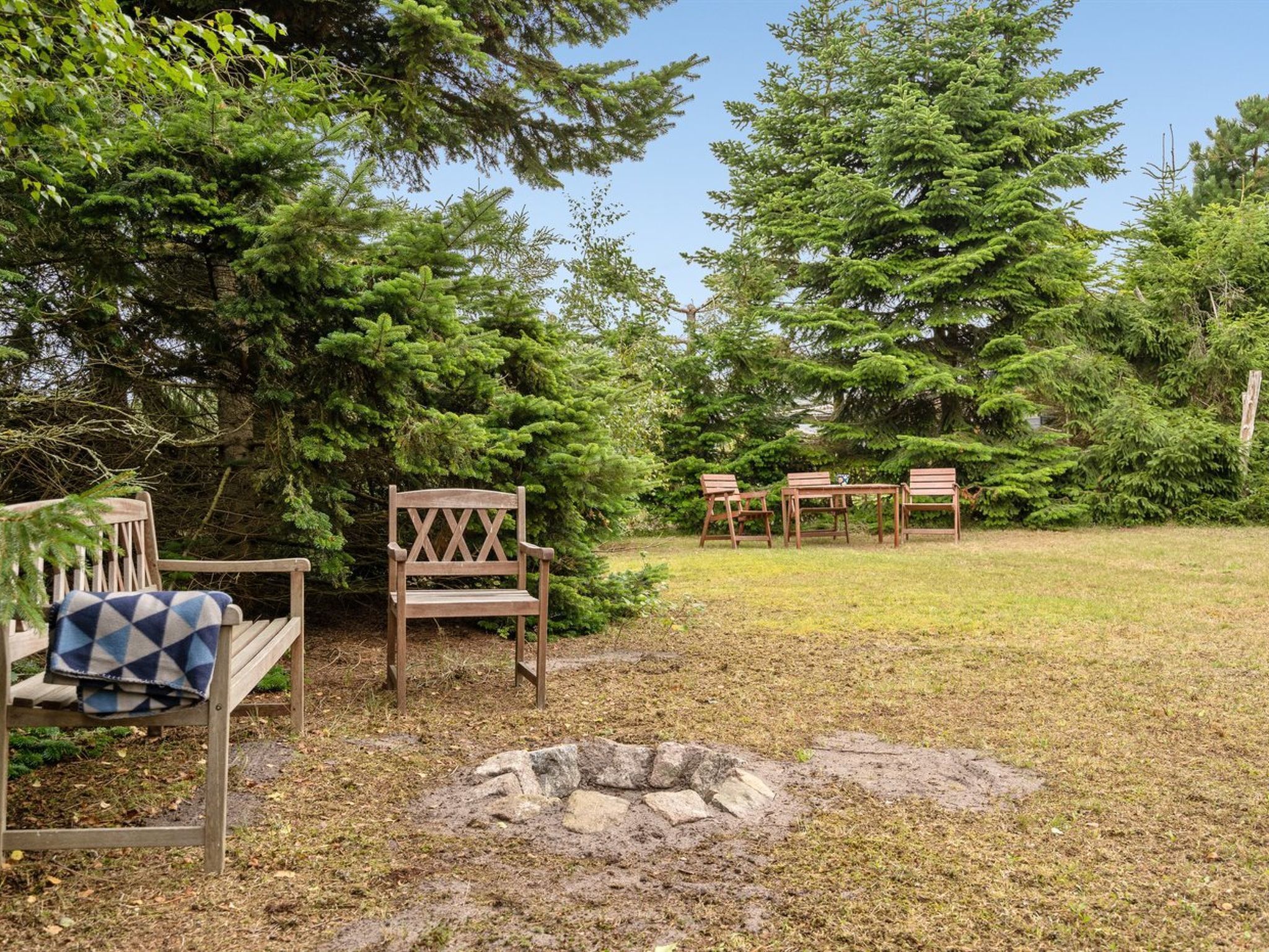 Un espace patio aménagé avec du mobilier en bois, notamment un banc et des tables, entouré d’une végétation luxuriante et d’arbres.