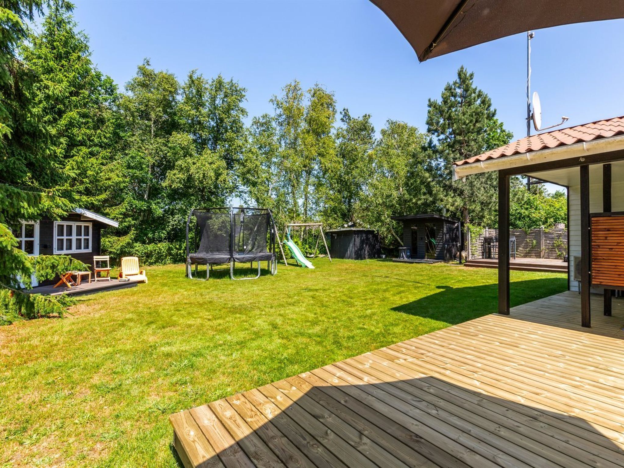 Un jardin avec un trampoline, un petit abri de jardin, une cabane et une terrasse en bois.