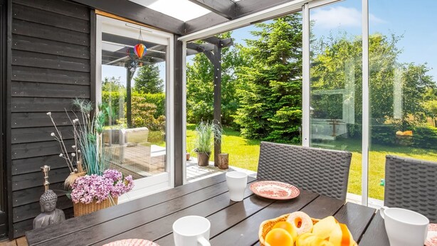 Un espace repas extérieur moderne, avec une table dressée pour deux personnes, entouré de chaises en osier, et offrant une vue sur un jardin luxuriant.