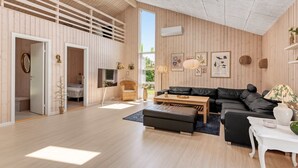 Un salon moderne avec un plancher en bois, un canapé noir, une table basse en bois et une petite table d’appoint blanche. Une grande fenêtre laisse entrer la lumière naturelle et offre une vue sur la verdure à l’extérieur.